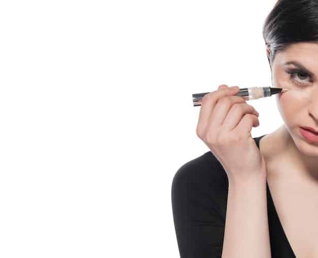 A woman demonstrating the tightlining technique on her upper eyelid, using a black waterproof eyeliner pencil to fill in the space between her lashes. Her hooded eye is partially open to show the subtle definition achieved.